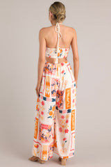 Back view of a patterned jumpsuit featuring a halter tassel tie neckline, an elastic waist, cutouts at the waist, functional pockets, and a smocked insert in the back of the bust.