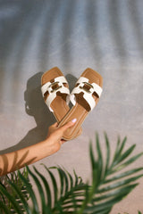 These white sandals feature a square toe, a slip-on design, and a strap with cutouts and a gold accent piece over the top of the foot.