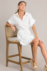 This white dress features a collared neckline, a partial button front, a self-tie drawstring waist, a raw hemline, and thick cuffed sleeves.