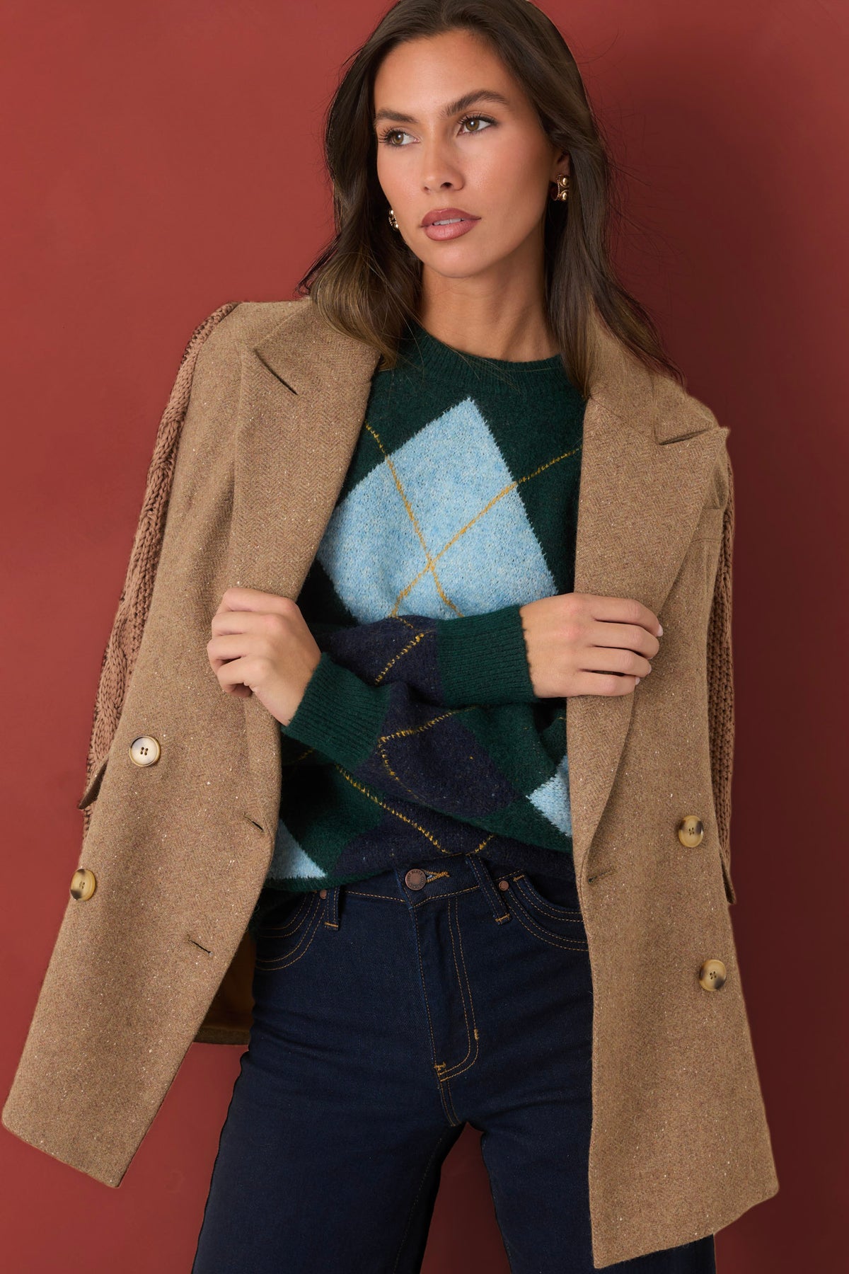 Woman modeling a forest green argyle sweater, a light brown knit blazer, and dark wash jeans in front of a burgundy backdrop. 