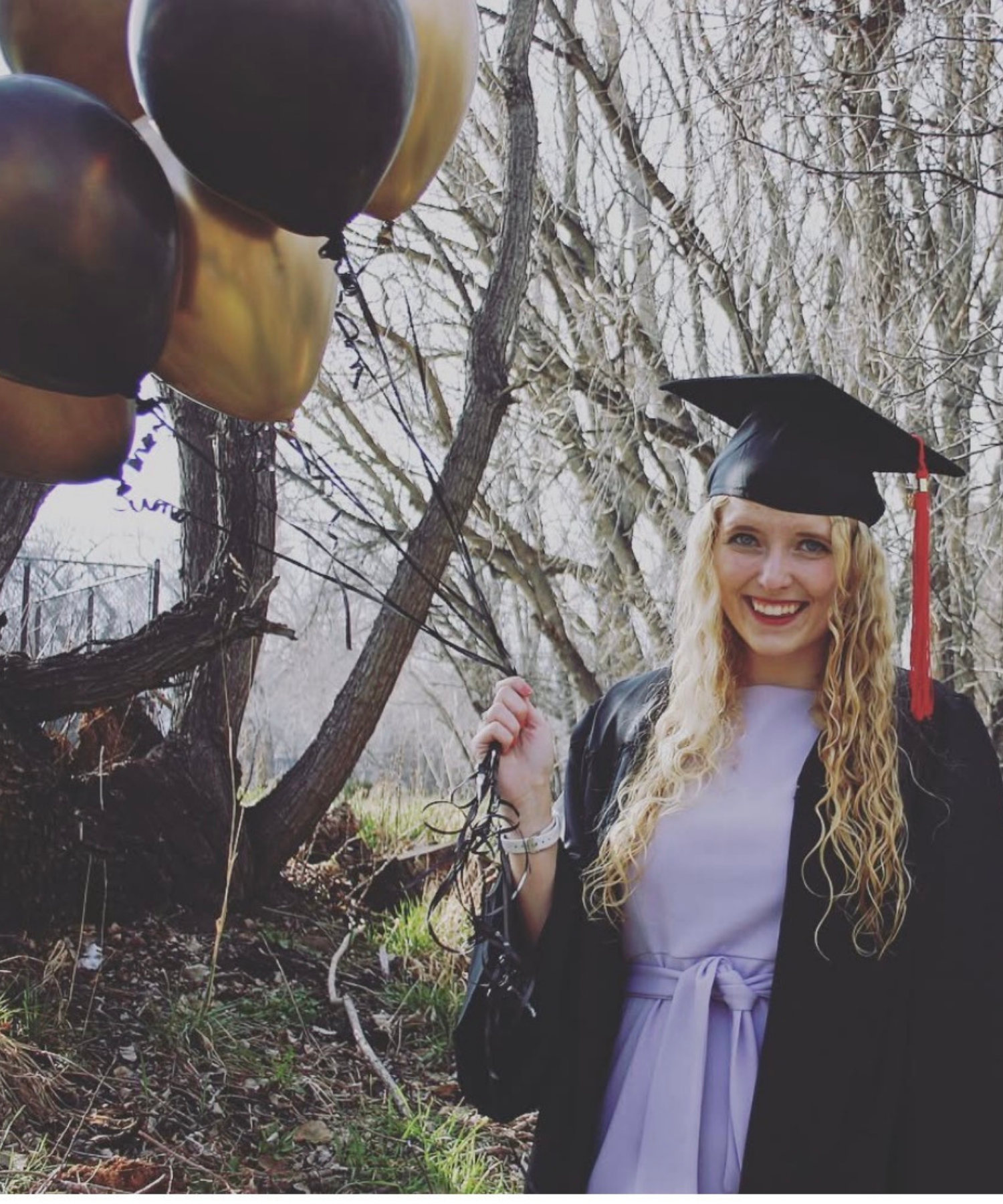Woman in a graduation cap and gown holding balloons outdoors with bare trees in the background.