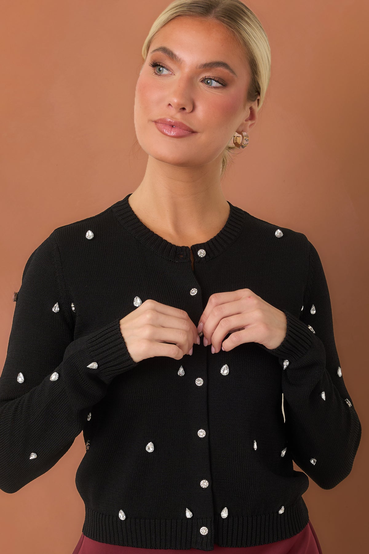 Woman modeling a black cardigan in front of a burnt orange backdrop. The cardigan showcases teardrop-shaped rhinestones scattered across the front and long sleeves.