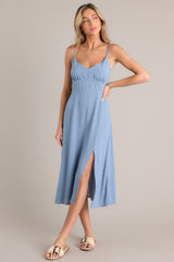 Angled full body view of this ash blue midi dress that features a v-neckline, thin adjustable straps, a smocked back insert, gathering in the bust, a thick waistband, and a side slit.