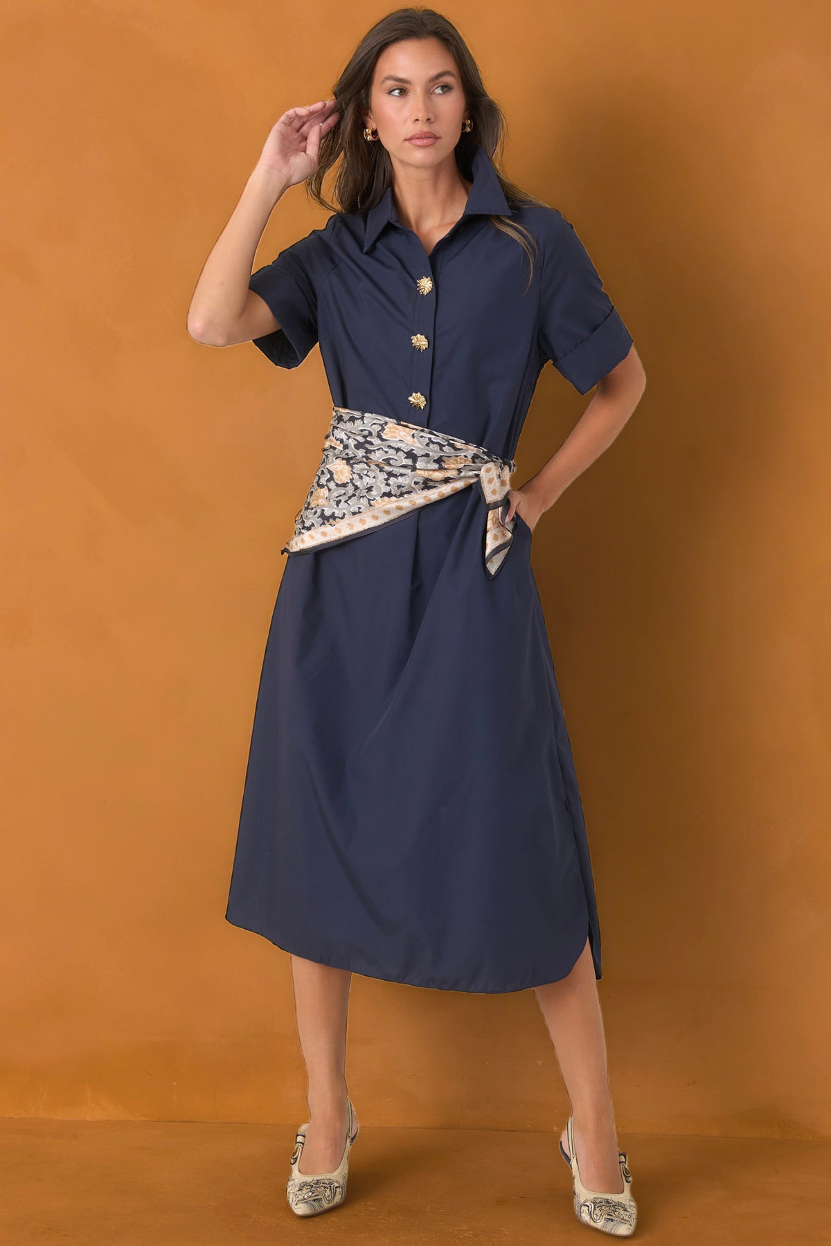 A woman models a navy blue short-sleeve midi shirt dress with gold buttons and a patterned cream and blue waist tie, set against a solid warm brown studio backdrop.