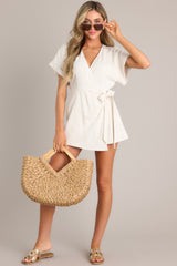 This beige romper features a V-shaped neckline, a keyhole back with button closure, cuffed short sleeves, and a faux wrap.