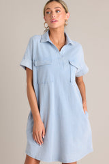 Angled front view of this light chambray mini dress that features a collared neckline, functional breast & hip pockets, a relaxed fit, and cuffed short sleeves.