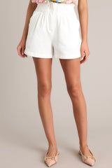 Front view of these ivory shorts that feature a high waisted paper bag design, an elastic waistband, a zipper closure, functional hip pockets, and faux back pockets.