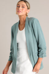 Angled front view of this forest green blazer with a folded neckline, shoulder padding, faux pockets, and gathered quarter-length sleeves.
