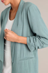 Close up view of this forest green blazer with a folded neckline, shoulder padding, faux pockets, and gathered quarter-length sleeves.