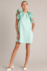 Full body view of this light green mini dress that features a high crew neckline, a unique floral pattern, functional hip pockets, and ruffled sleeves.