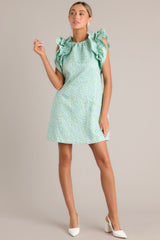 In a soft light green shade, this mini dress boasts a high crew neckline, a distinctive floral pattern, practical hip pockets, and charming ruffled sleeves