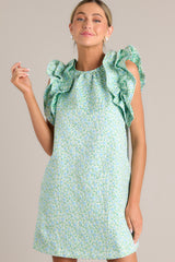 Close up front view of this light green mini dress that features a high crew neckline, a unique floral pattern, functional hip pockets, and ruffled sleeves.
