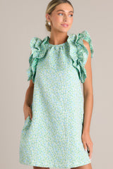 Angled close up view of this light green mini dress that features a high crew neckline, a unique floral pattern, functional hip pockets, and ruffled sleeves.