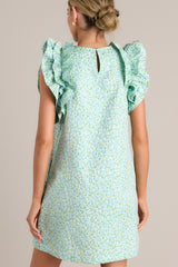 Back view of this light green mini dress that features a high crew neckline, a unique floral pattern, functional hip pockets, and ruffled sleeves.