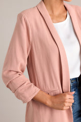 Close up view of this dusty rose blazer featuring a folded neckline, shoulder padding, faux pockets, and gathering in the quarter-length sleeves.