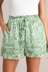 Close up view of these green tropical print shorts that feature a high waisted design, an elastic ruffled waistband, a self-tie drawstring, functional hip pockets, and a wide leg.