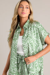This green tropical print top features a collared neckline, a functional button front, wide folded short sleeves, and a split hemline.