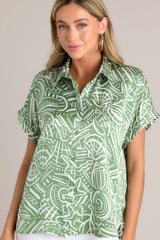 Front view of this green tropical print top that features a collared neckline, a functional button front, wide folded short sleeves, and a split hemline.