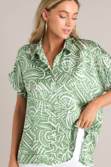 Angled close up view of this green tropical print top that features a collared neckline, a functional button front, wide folded short sleeves, and a split hemline.