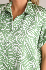 Close up view of this green tropical print top that features a collared neckline, a functional button front, wide folded short sleeves, and a split hemline.