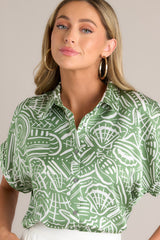 Close up front view of this green tropical print top that features a collared neckline, a functional button front, wide folded short sleeves, and a split hemline.