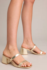 Angled side view of these gold block heels that feature a square toe, two gold straps across the top of foot, and a block heel.