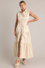Front view of  this beige maxi dress that features a collared neckline, a full button front, buttoned breast pockets, a thick elastic waistband, buttoned hip pockets, and a tiered design.