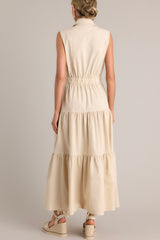 Back view of this beige maxi dress that features a collared neckline, a full button front, buttoned breast pockets, a thick elastic waistband, buttoned hip pockets, and a tiered design.