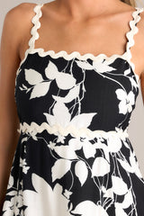 Close up view of this black floral midi dress that features a square neckline, thin ricrac straps, a fully smocked back, ricrac detailing, functional hip pockets, and a ricrac hemline.
