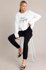 Seated full body view of this white sweater that features a ribbed crew neckline, embroidered wording, a soft knitted fabric, and a ribbed hemline.