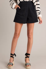 Front view of these black shorts feature a high waisted design, elastic insert in the back of the waist, hook & bar closure, an added button for extra security, a zipper, belt loops, functional hip pockets, and pleated details.
