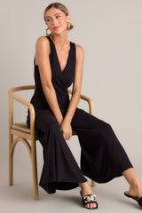 Seated full body view of this black jumpsuit features a v-neckline, a wrap-style bodice, a fitted waist, functional hip pockets, and an extremely wide & flowing leg.