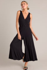 Full body view of this black jumpsuit features a v-neckline, a wrap-style bodice, a fitted waist, functional hip pockets, and an extremely wide & flowing leg.