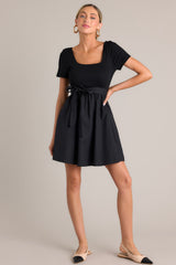 This black mini dress features a square neckline, a ribbed bodice, an elastic waistband, belt loops, a self-tie waist belt, and a flowing silhouette.