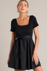 Front view of this black mini dress that features a square neckline, a ribbed bodice, an elastic waistband, belt loops, a self-tie waist belt, and a flowing silhouette.