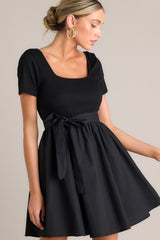 Angled front view of this black mini dress that features a square neckline, a ribbed bodice, an elastic waistband, belt loops, a self-tie waist belt, and a flowing silhouette.
