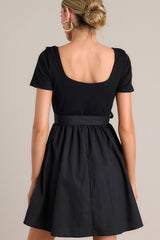 Back view of this black mini dress that features a square neckline, a ribbed bodice, an elastic waistband, belt loops, a self-tie waist belt, and a flowing silhouette.