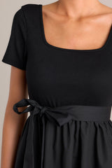 Close up view of this black mini dress that features a square neckline, a ribbed bodice, an elastic waistband, belt loops, a self-tie waist belt, and a flowing silhouette.