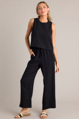 Full body view of these black pants that feature a high waisted design, an elastic waistband, a self-tie drawstring, functional hip pockets, and a wide leg.