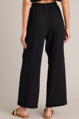 Back view of these black pants that feature a high waisted design, an elastic waistband, a self-tie drawstring, functional hip pockets, and a wide leg.