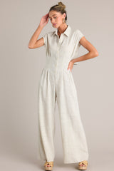 Full body view of this beige jumpsuit that features a collared neckline, a functional button front, a cinched waist, an elastic insert in the back of the waist, a lower back cutout, functional pockets, and a wide leg.