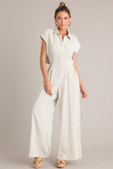 Front view of this beige jumpsuit that features a collared neckline, a functional button front, a cinched waist, an elastic insert in the back of the waist, a lower back cutout, functional pockets, and a wide leg.