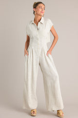 Angled front view of this beige jumpsuit that features a collared neckline, a functional button front, a cinched waist, an elastic insert in the back of the waist, a lower back cutout, functional pockets, and a wide leg.