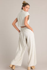 Back view of this beige jumpsuit that features a collared neckline, a functional button front, a cinched waist, an elastic insert in the back of the waist, a lower back cutout, functional pockets, and a wide leg.