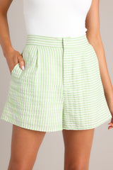 Close up view of these light green high-waisted shorts with elastic insert at back waist, hook & bar closure, functional zipper, pleat detailing, hip pockets, striped pattern.
