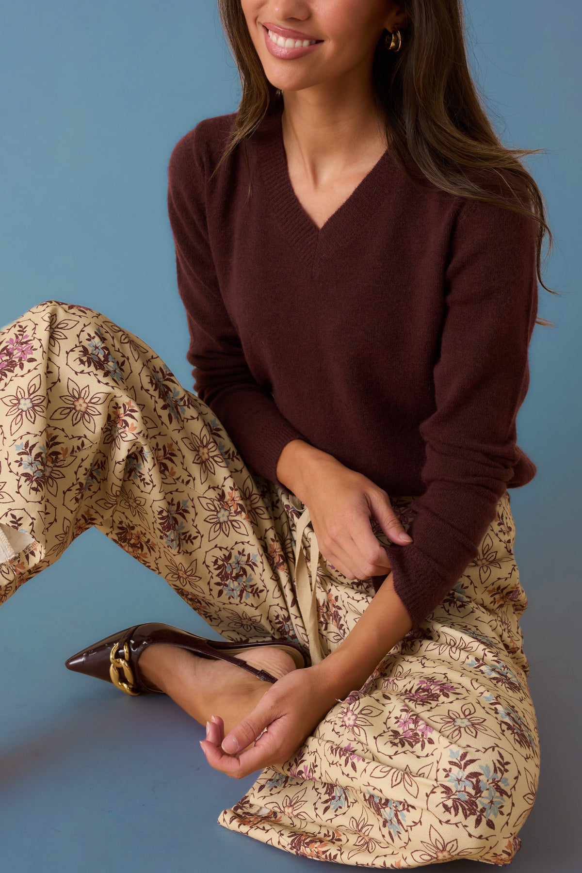 Woman in a burgundy sweater and beige floral print pants against a faded blue backdrop. 