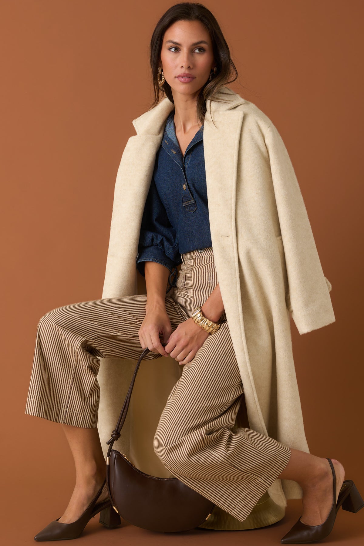 Woman in an oatmeal trench coat, denim top, and brown striped pants against a sienna backdrop. 