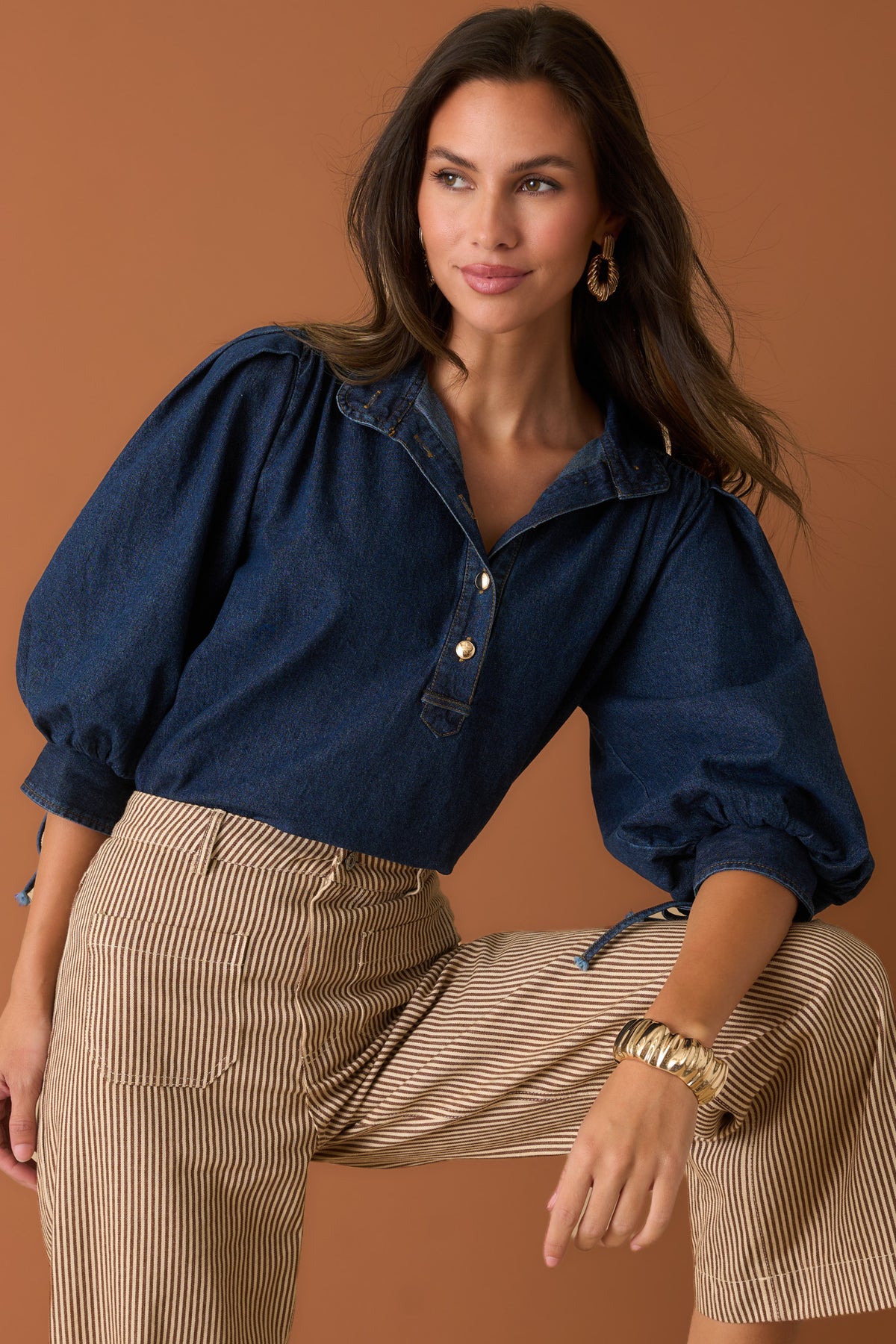 Woman in a denim, balloon sleeve top, paired with brown striped pants against a sienna backdrop.