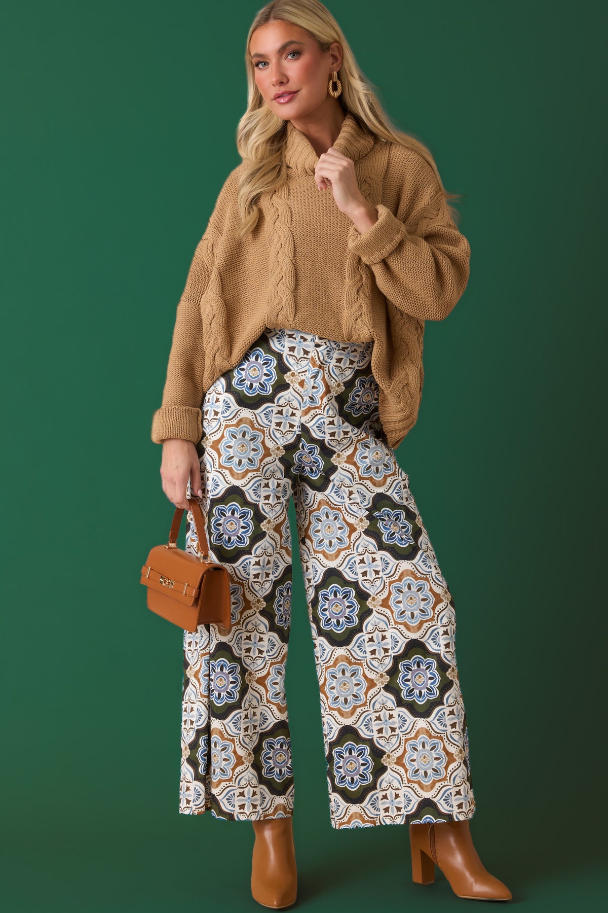 Woman wearing olive, ivory, and brown geometric print pants with a camel cable knit sweater against a forest green backdrop.