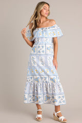 Full front view of this white & blue maxi dress featuring an elastic off-the-shoulder neckline, a frilled overlay that forms a false sleeve, an elastic stretch waist, an embroidered floral design, and a lined and tiered skirt.
